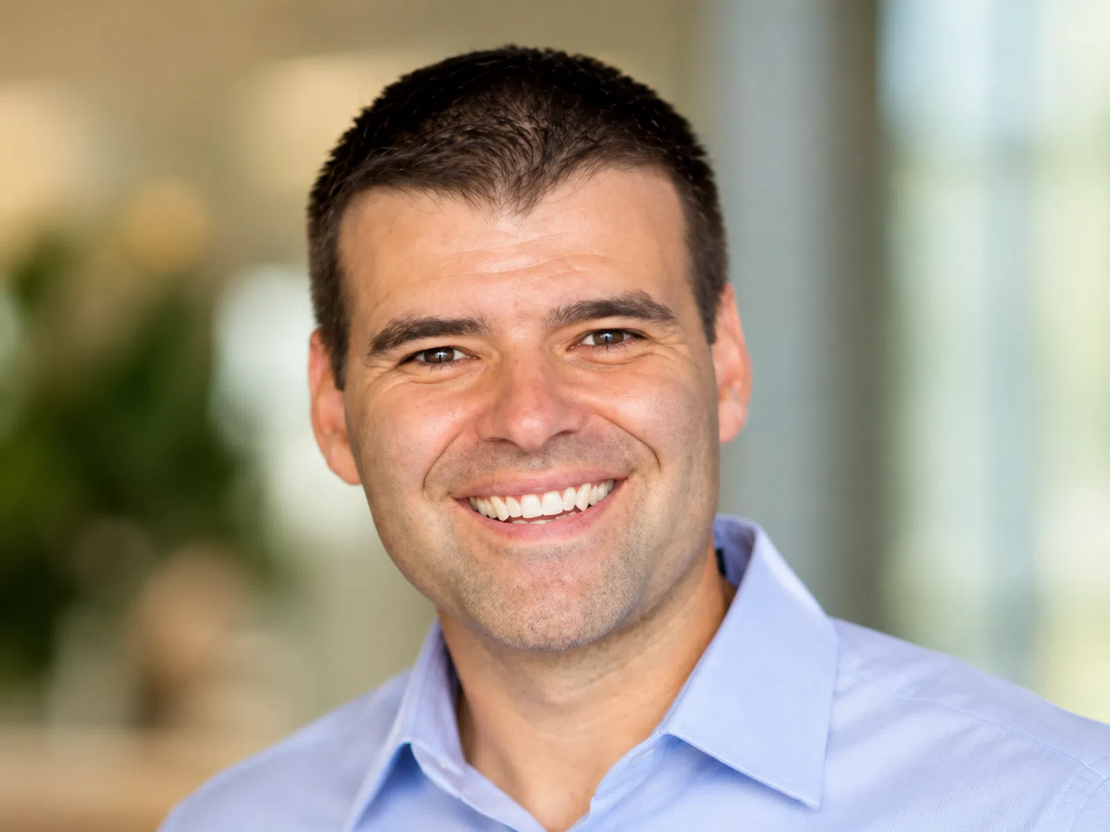 Smiling man with short dark hair wearing a light blue button-up shirt against a blurred background.