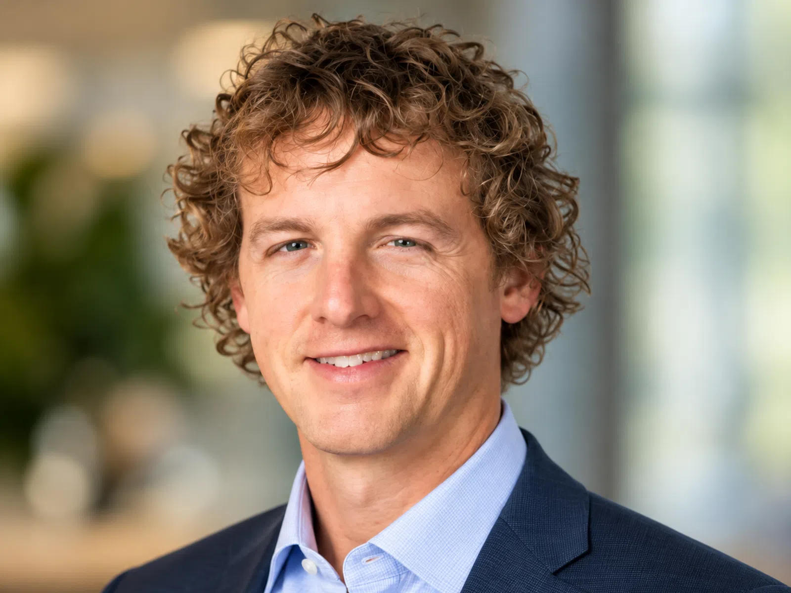 Smiling man with curly hair wearing a blue suit jacket and light blue shirt in a blurred office background.