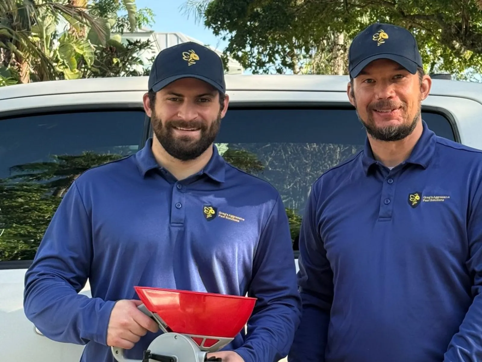 Two men in blue uniforms with company logos stand by a white truck, holding a red tool under a sunny tree.