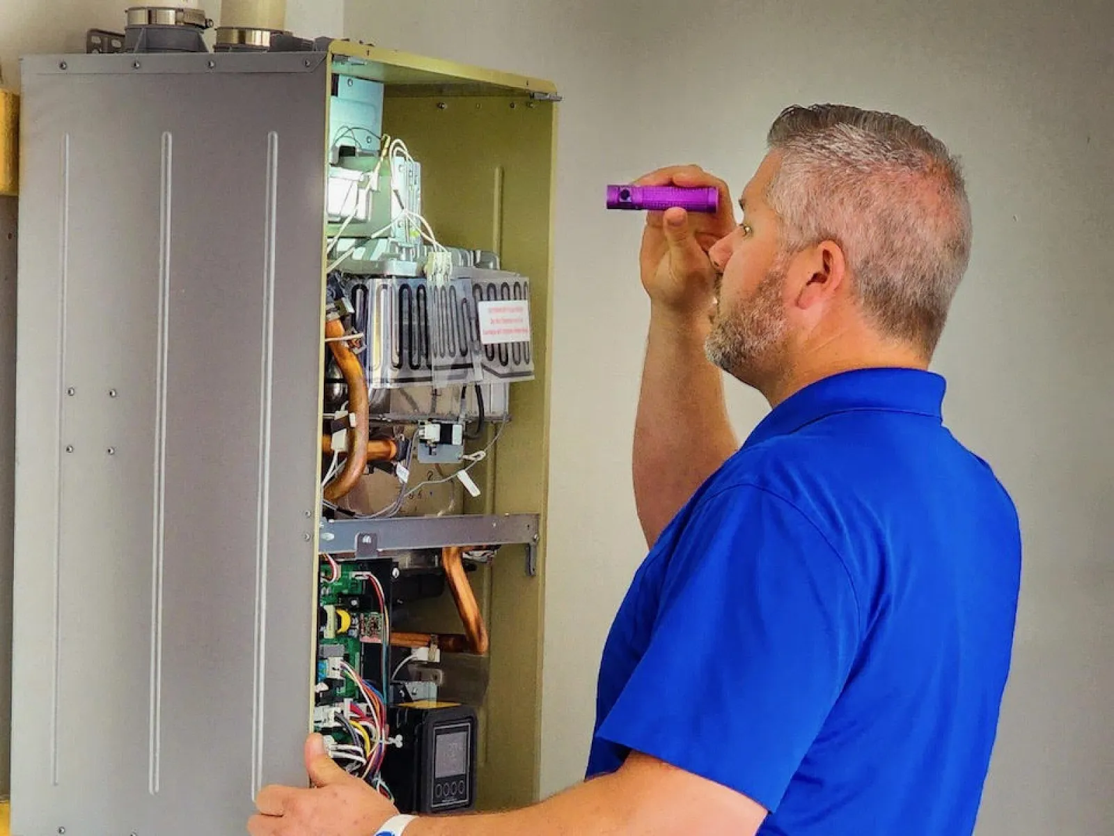 Technician in blue shirt inspects a wall-mounted heating unit with a flashlight during maintenance work.
