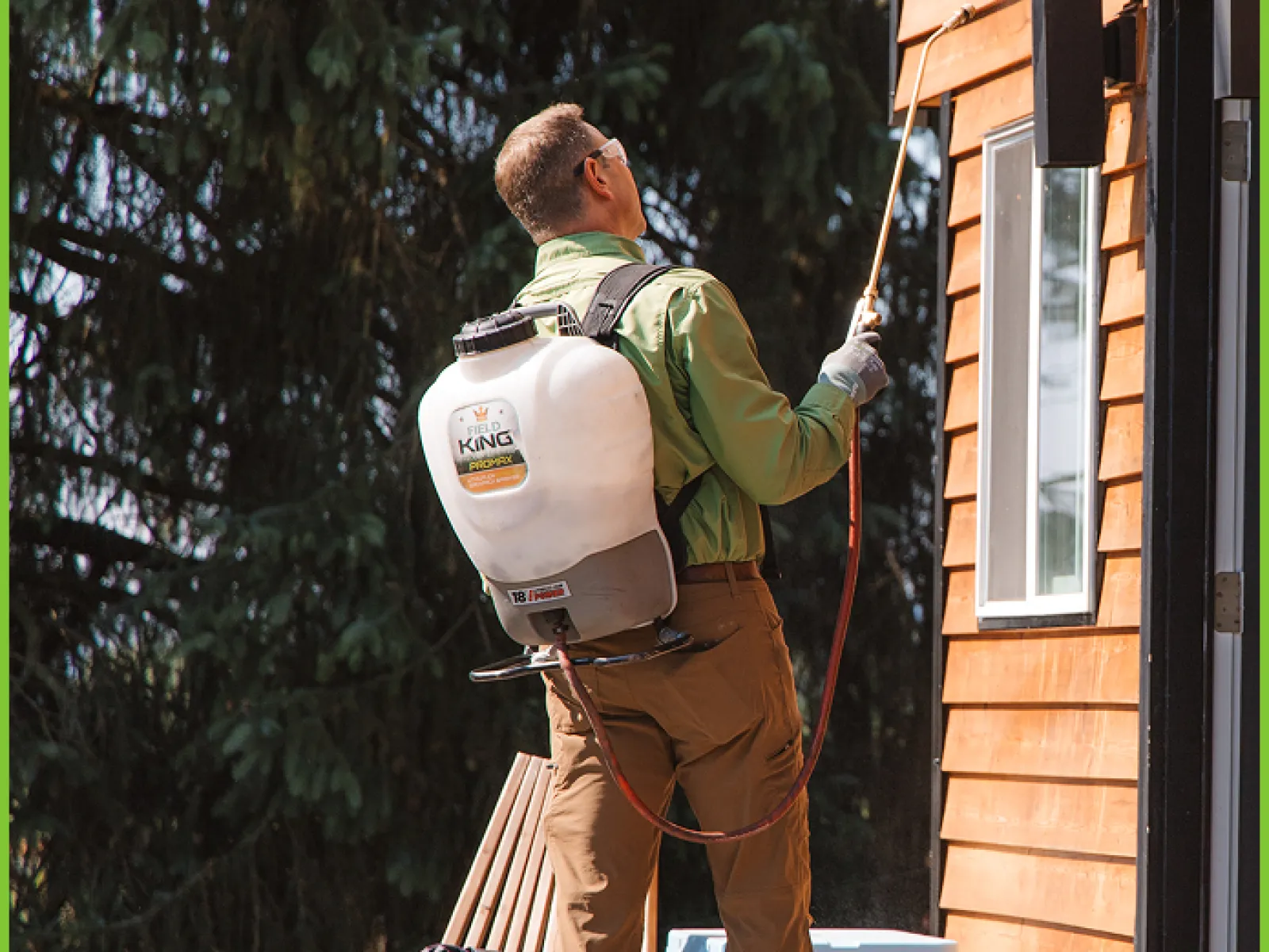 Man wearing green shirt spraying exterior wooden wall of house with backpack sprayer in daylight.