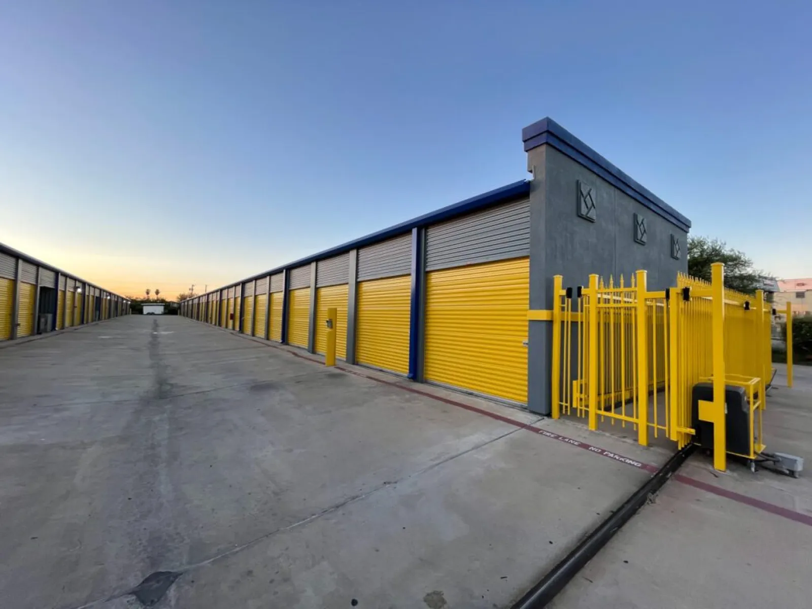 Outdoor storage facility with yellow roll-up doors and gated entrance under clear sky at dusk.