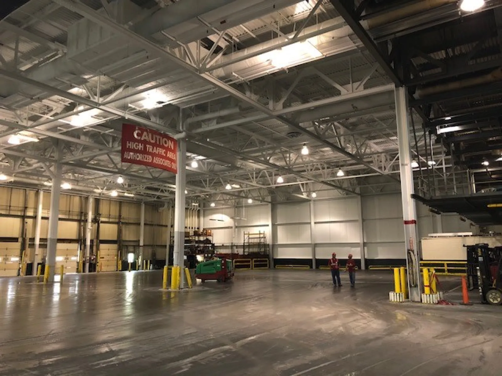 Spacious industrial warehouse interior with high ceilings, bright lighting, and two workers near equipment.