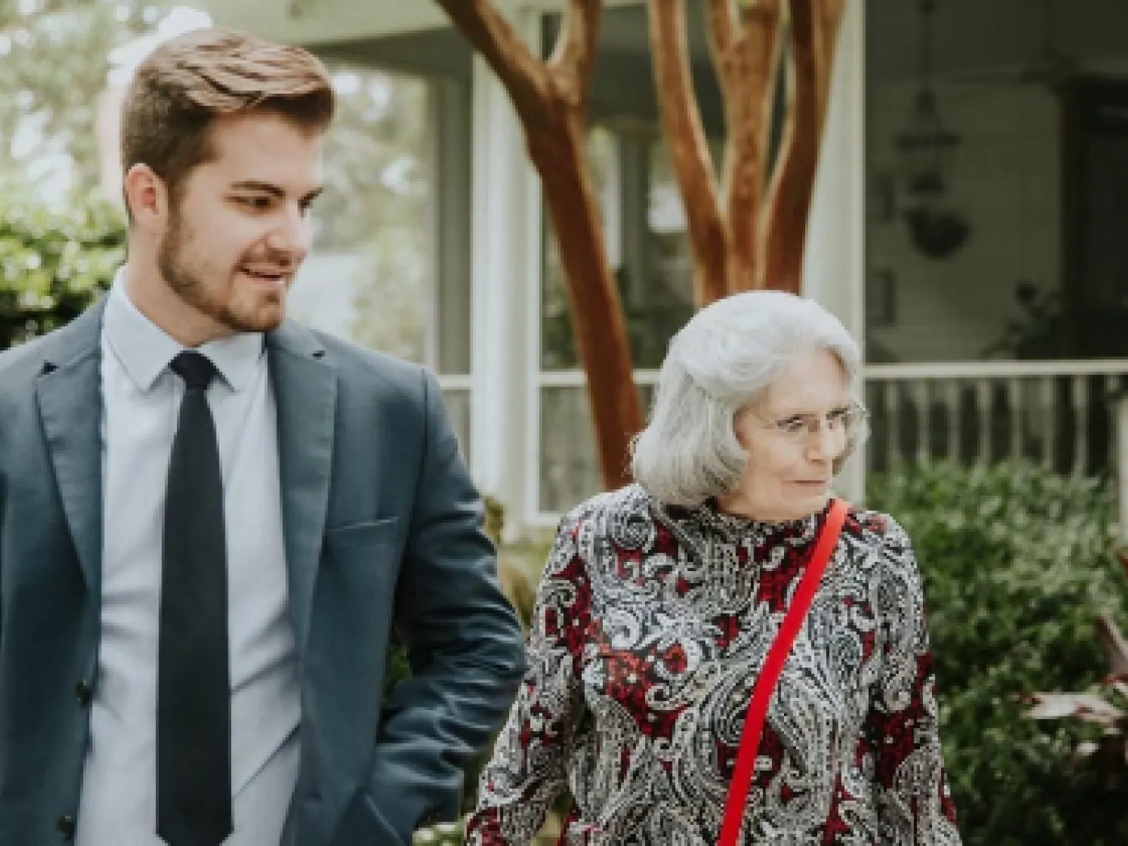 Young man in suit walks outdoors with elderly woman wearing patterned dress and glasses in a garden setting.