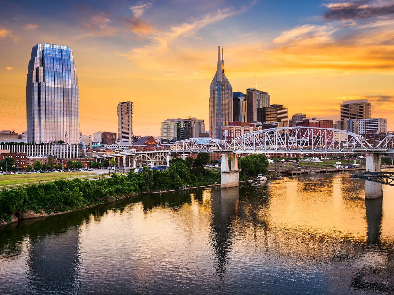 Nashville skyline at sunset with Cumberland River and John Seigenthaler Pedestrian Bridge reflecting in water.