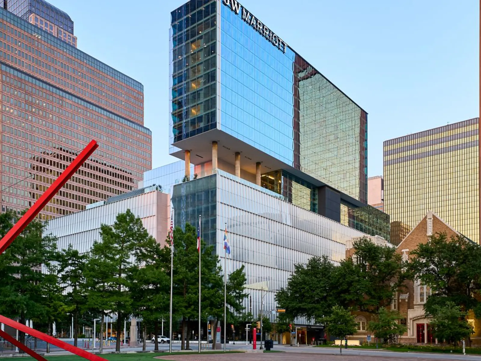 Cityscape featuring modern buildings, green trees, and a large red sculpture in the foreground.
