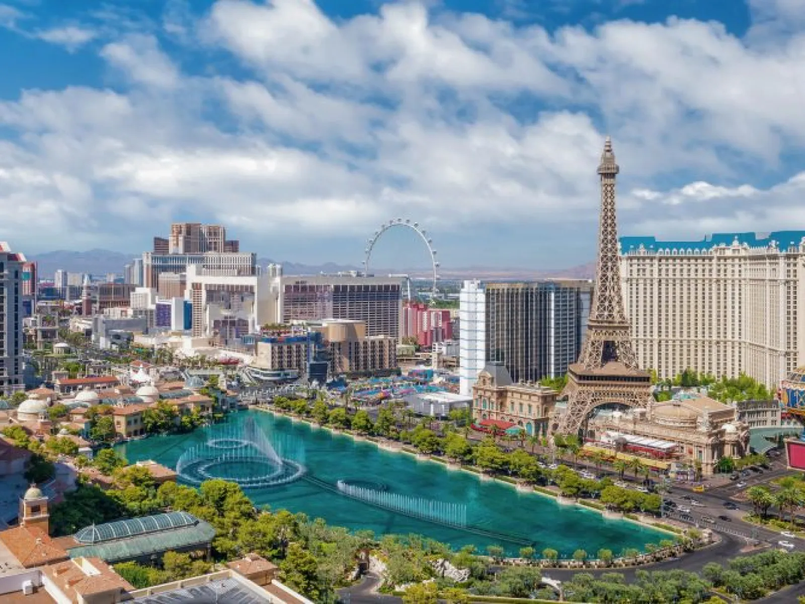 Aerial view of Las Vegas Strip showing hotels, Eiffel Tower replica, fountains, and high-rise buildings under blue sky.