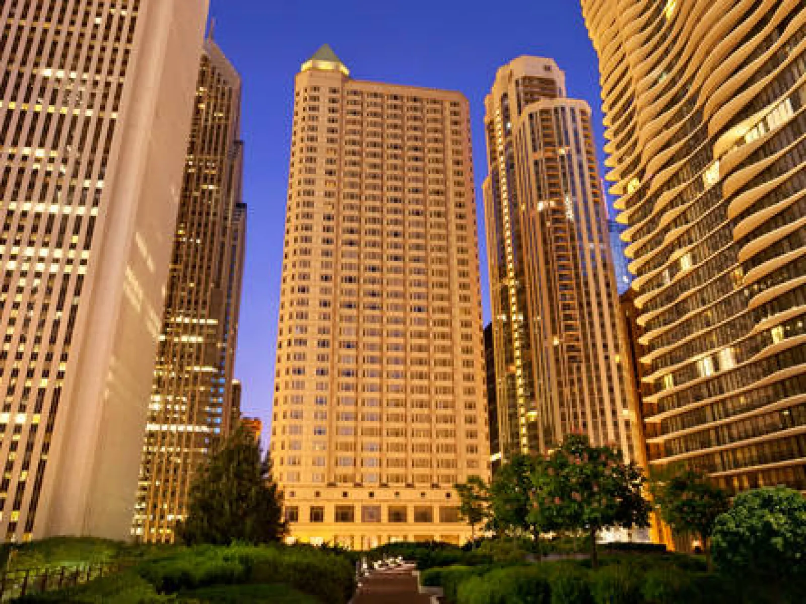 Modern skyscrapers with glass facades at sunset in a city park setting with green trees and clear sky.