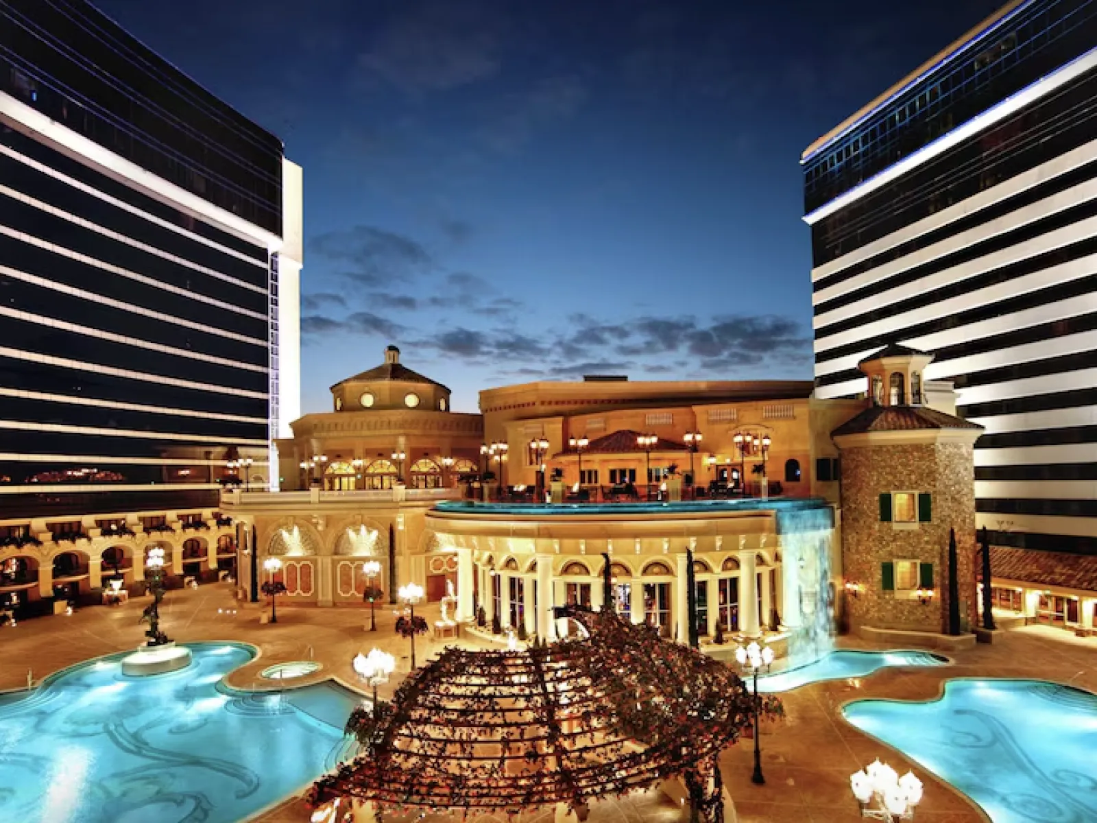Luxury hotel courtyard at dusk with illuminated pool, waterfall, and arched architecture between modern buildings.
