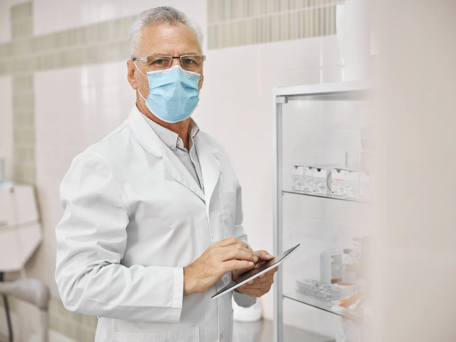 Senior male doctor in lab coat and face mask using tablet in medical clinic or hospital setting.