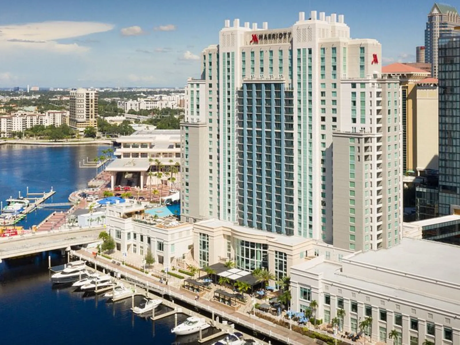 Tall Marriott hotel building overlooking a waterfront marina with boats and a city skyline in the background.
