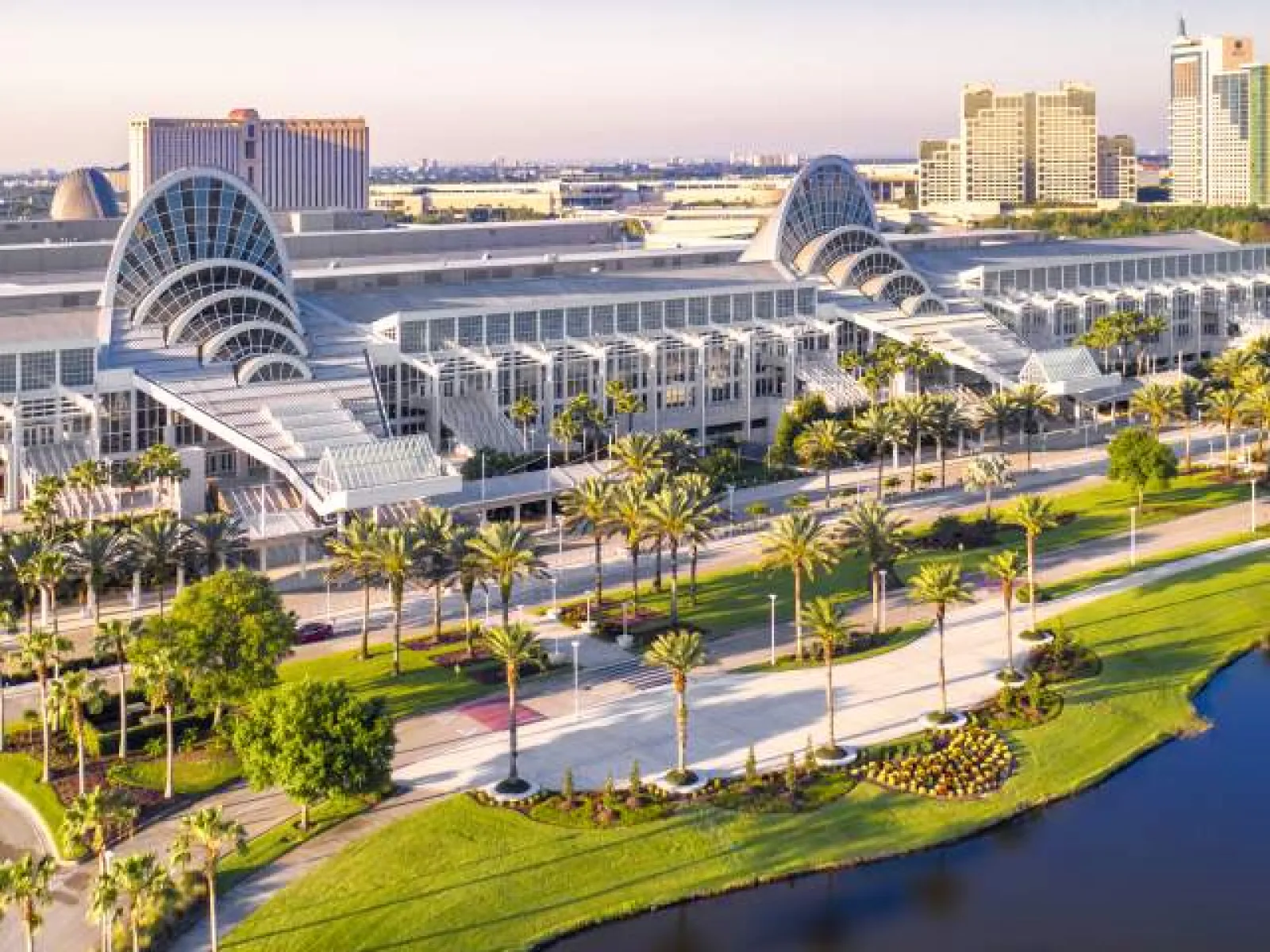 Convention center with unique arched glass structures, palm trees, green lawn, and adjacent water body under clear sky.