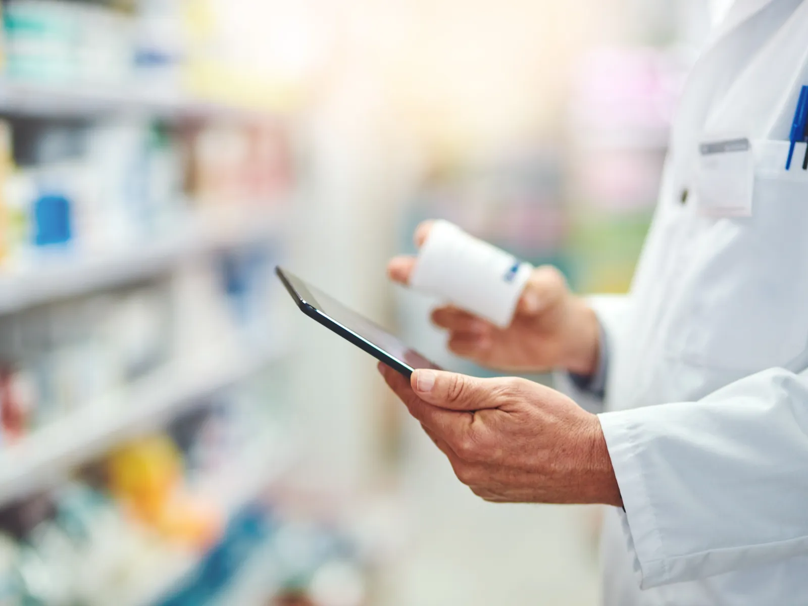 Pharmacist in white coat checking medication details using digital tablet inside pharmacy aisle.