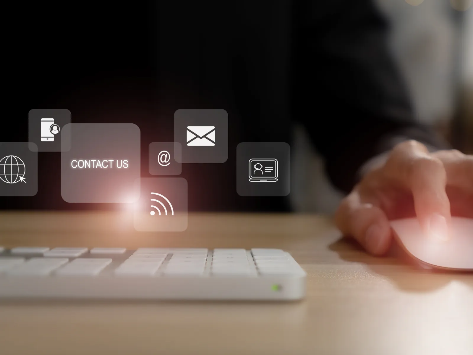 Hand using mouse with keyboard and digital contact us icons floating over a wooden desk in a dark setting