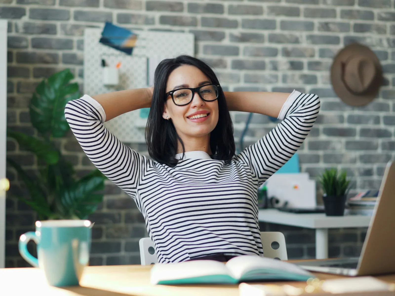 Smiling woman in glasses relaxing at desk with book, laptop, and plants in a cozy modern home office.
