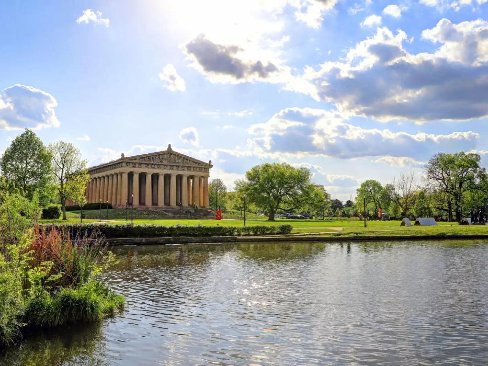 Parthenon replica in Nashville park reflected in calm water with blue sky and green trees surrounding it