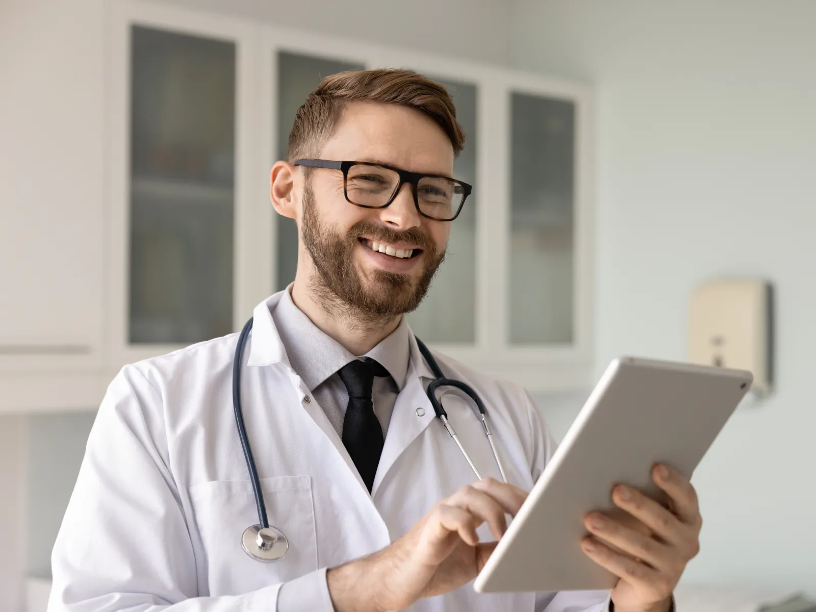Smiling male doctor wearing white coat and glasses using a digital tablet in a clinical setting.