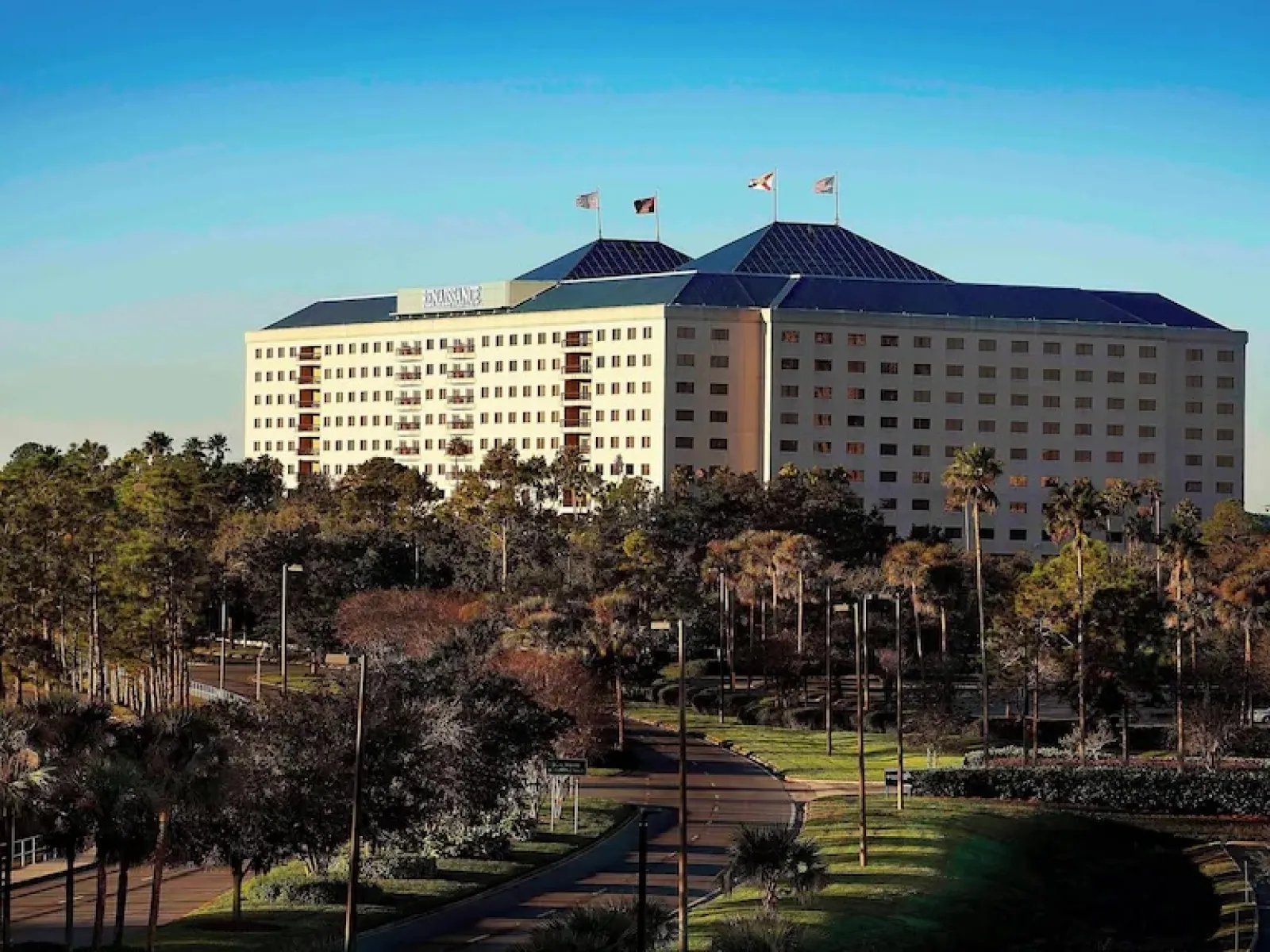 Large hotel building with multiple windows and flags on the roof surrounded by trees and a winding road under clear blue sky