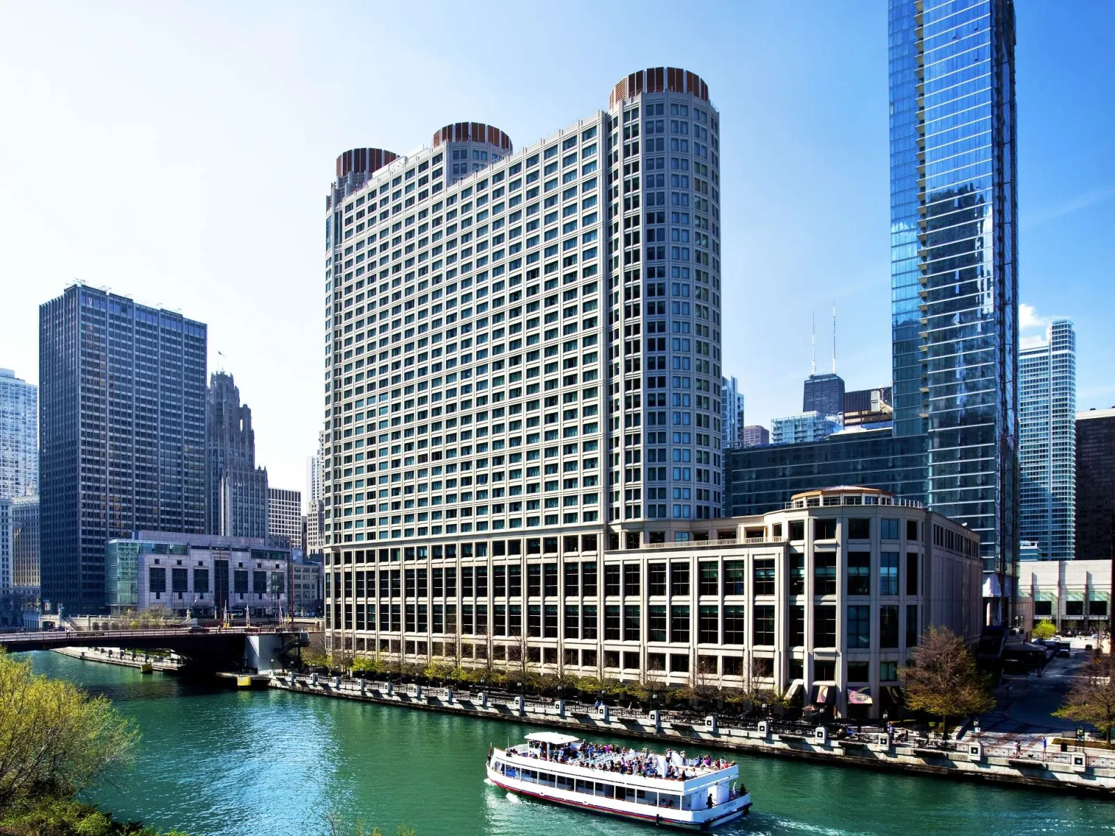 Tour boat cruising on river beside modern high-rise buildings in a sunny cityscape with clear blue sky.