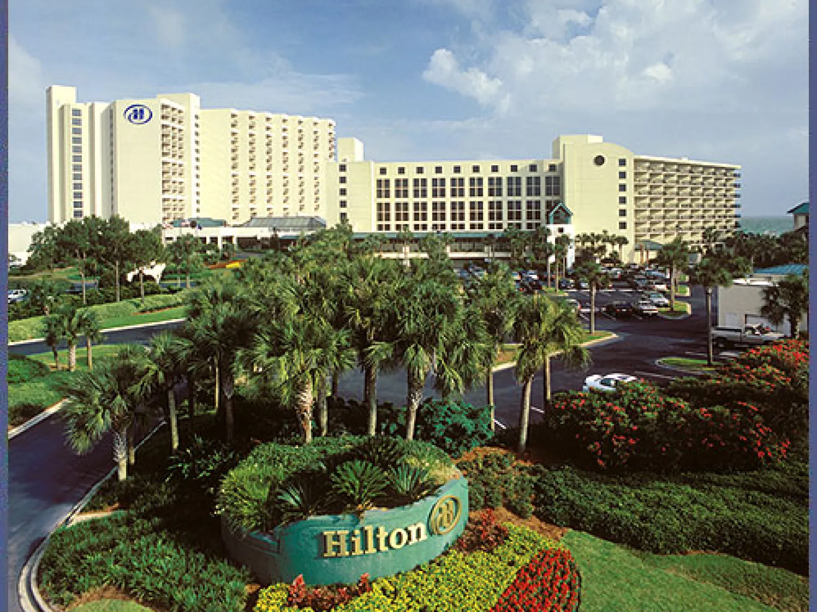 Hilton hotel with tall building, palm trees, lush gardens, and parking lot under a partly cloudy sky.