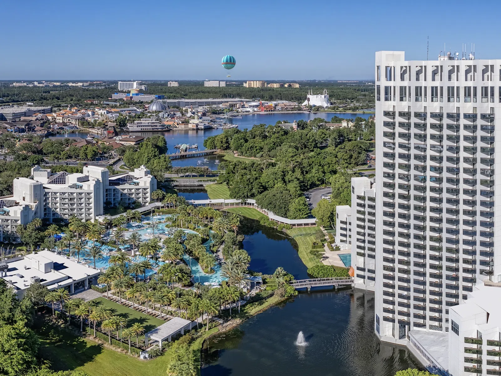Aerial view of Orlando resort with hotel towers, pools, green areas, water features, and a hot air balloon in the sky.