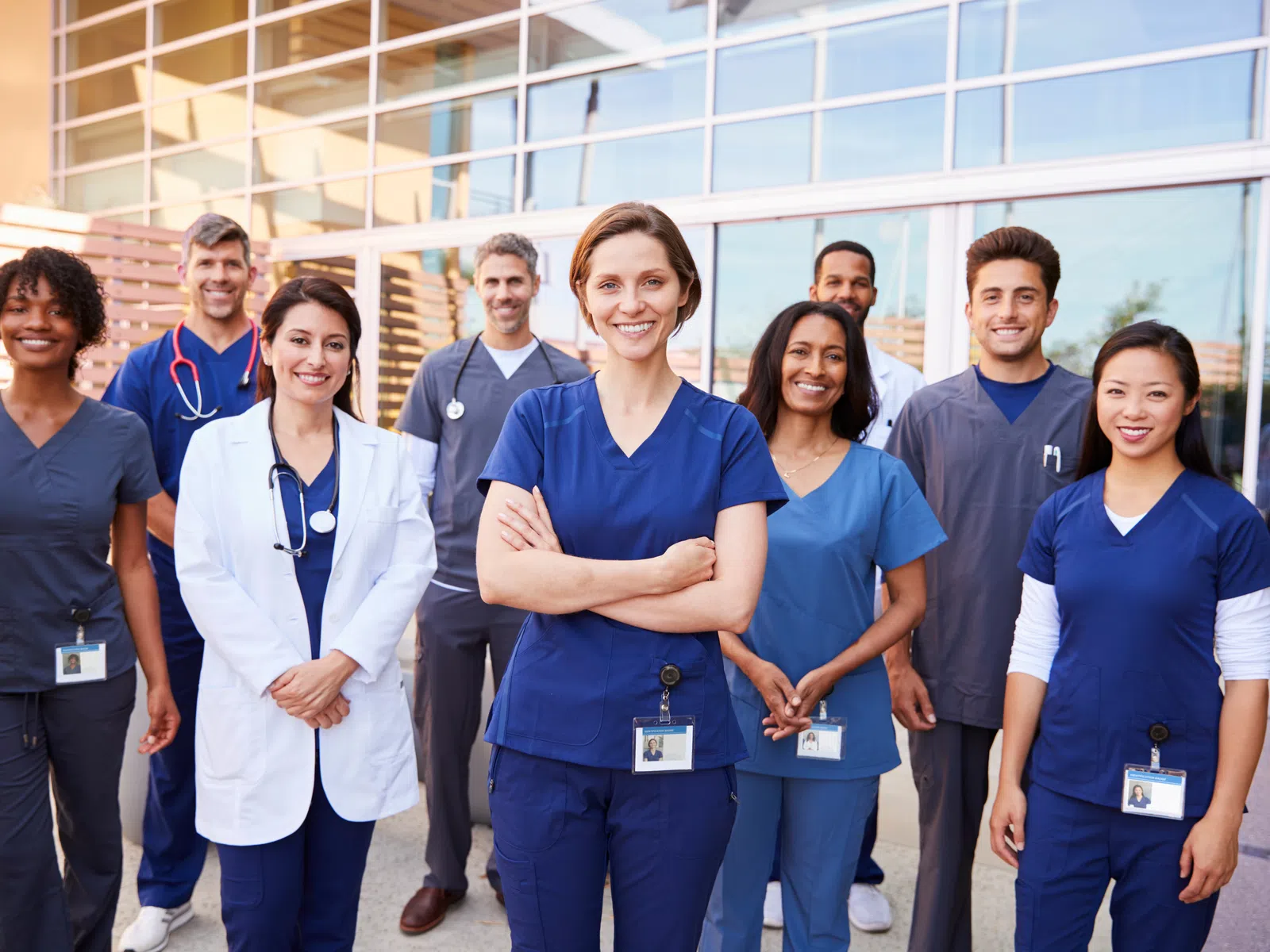 Diverse group of smiling medical professionals in scrubs and lab coats standing outside a modern hospital building.