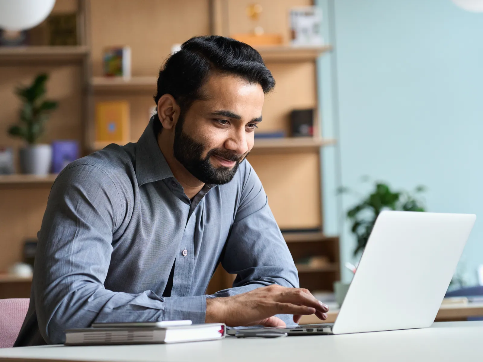 A smiling man working on a laptop in a modern workspace filled with greenery and books.