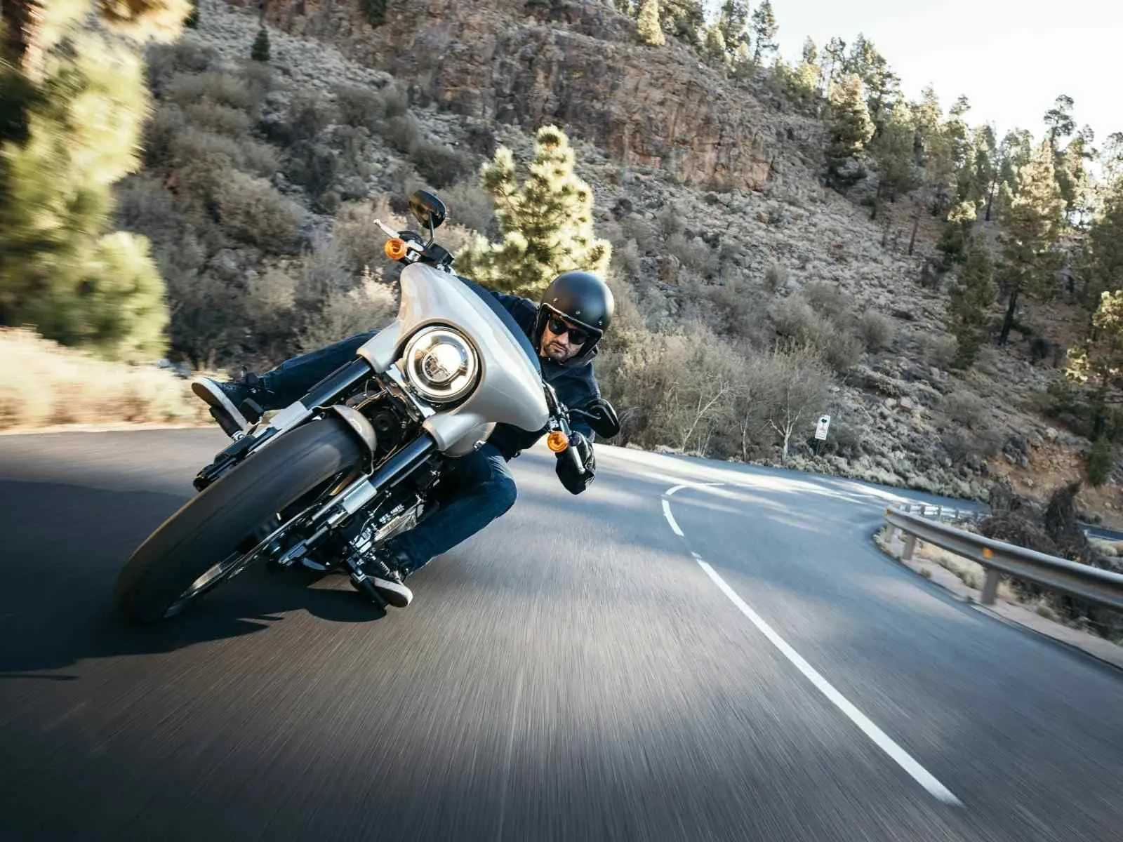 Motorcyclist wearing helmet leaning into a turn on a winding mountain road surrounded by trees and rocks