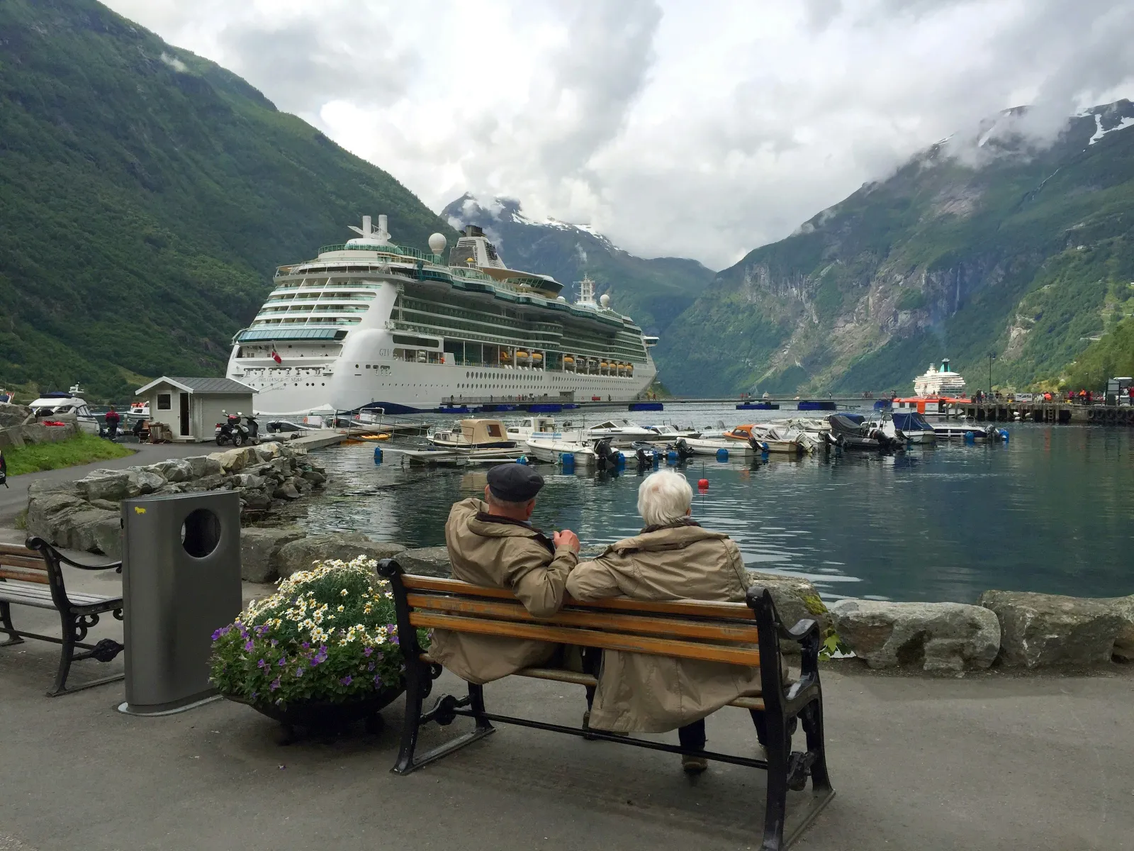 Elderly couple sitting on bench overlooking cruise ship docked in scenic fjord with mountains and cloudy sky