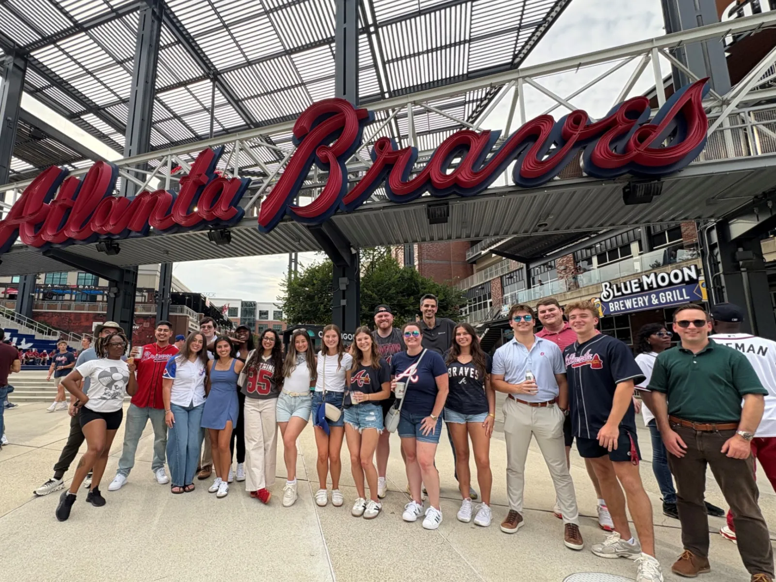 Group of people posing under the large Atlanta Braves sign outside the stadium on a cloudy day.