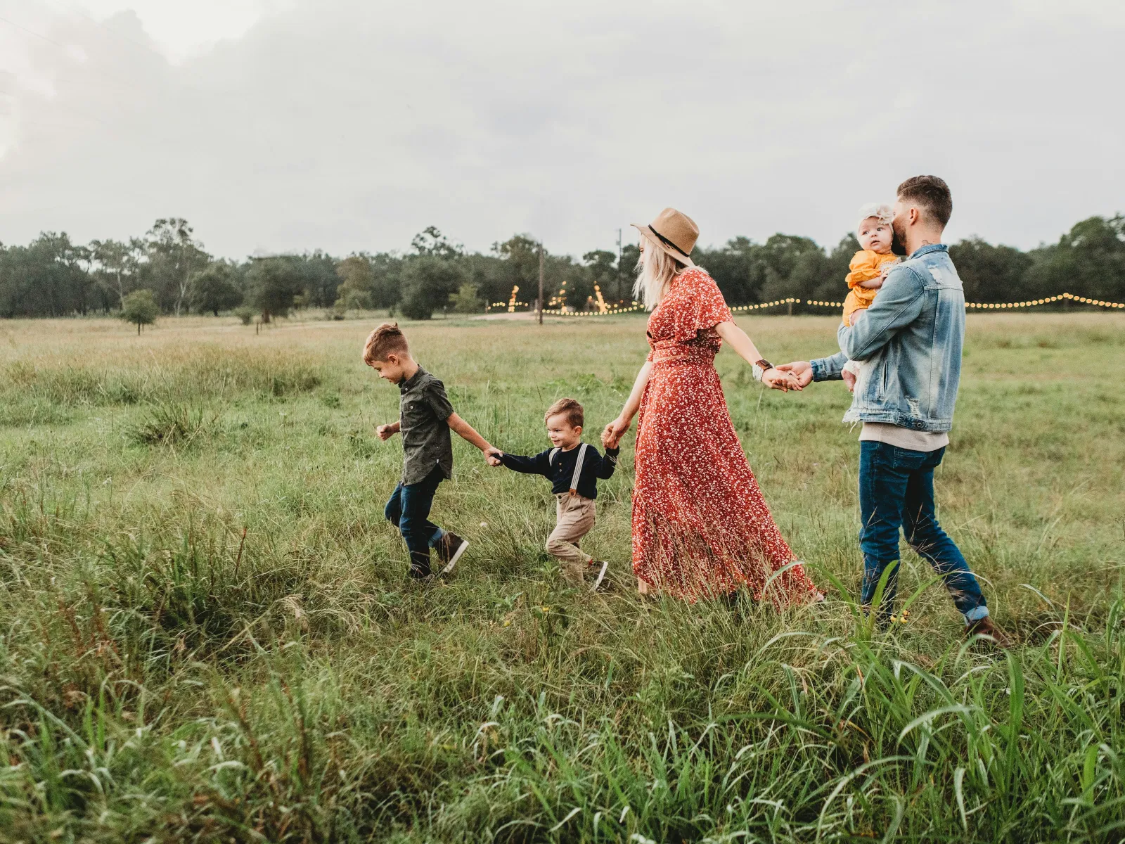 Family walking hand in hand through a grassy field with string lights in the background during daytime.