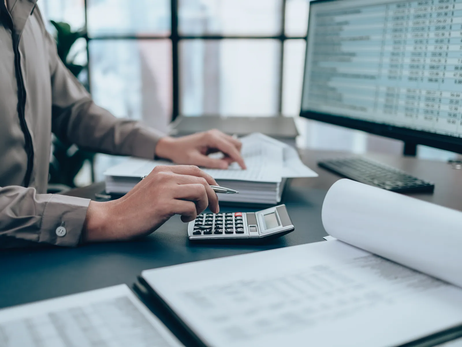 Person using calculator and analyzing financial documents at a desk with computer monitor showing data.