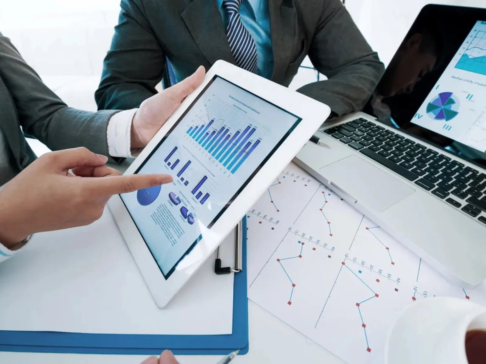 Business meeting with people analyzing data charts on tablet and laptop with printed graphs on table.