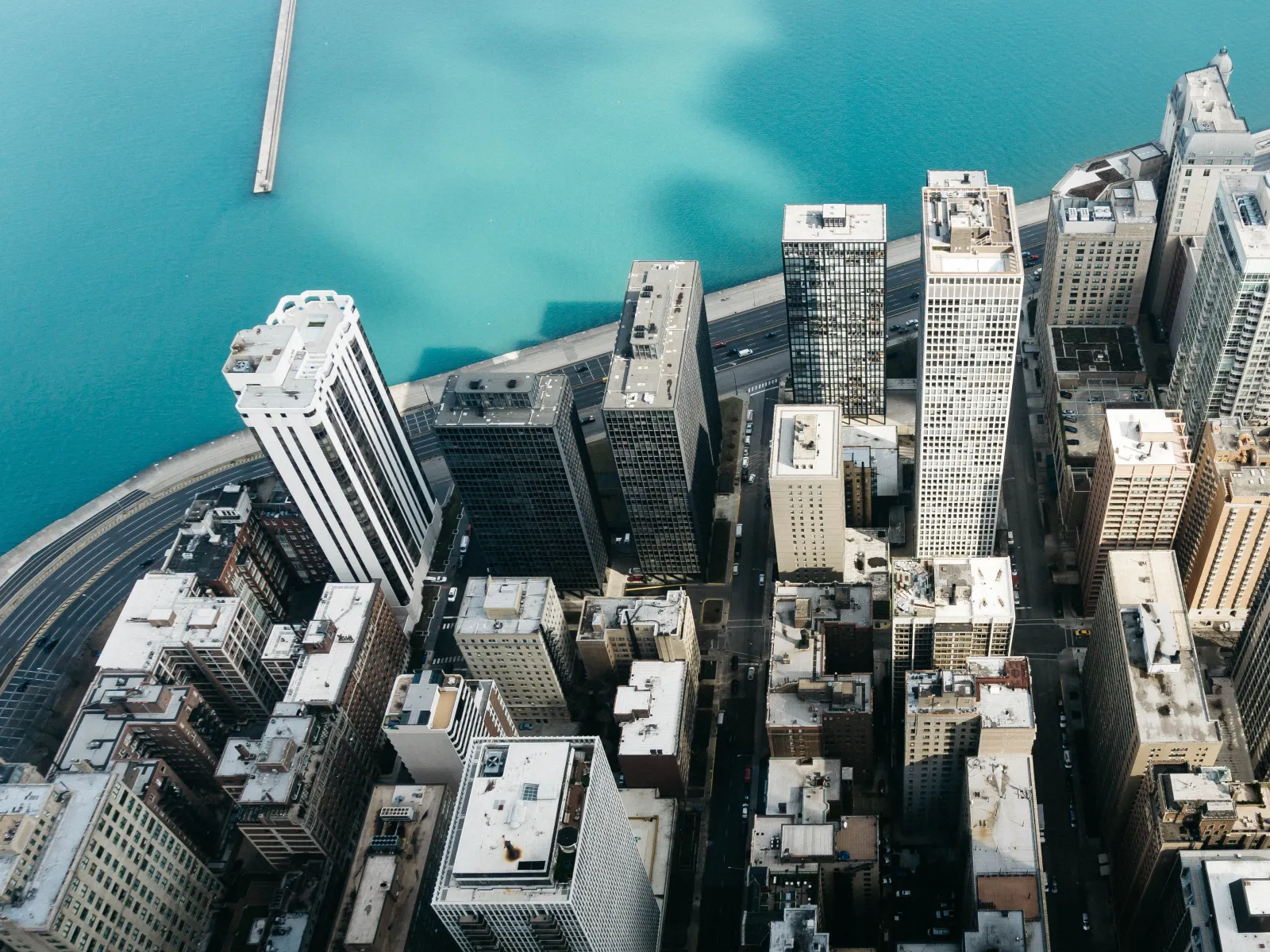 Aerial view of city skyscrapers by a blue waterfront with a pier extending into the water on a clear day.