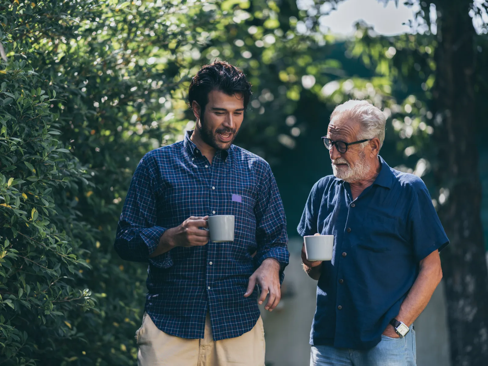 Two men, one young and one older, enjoy coffee while walking outdoors surrounded by greenery and sunlight.