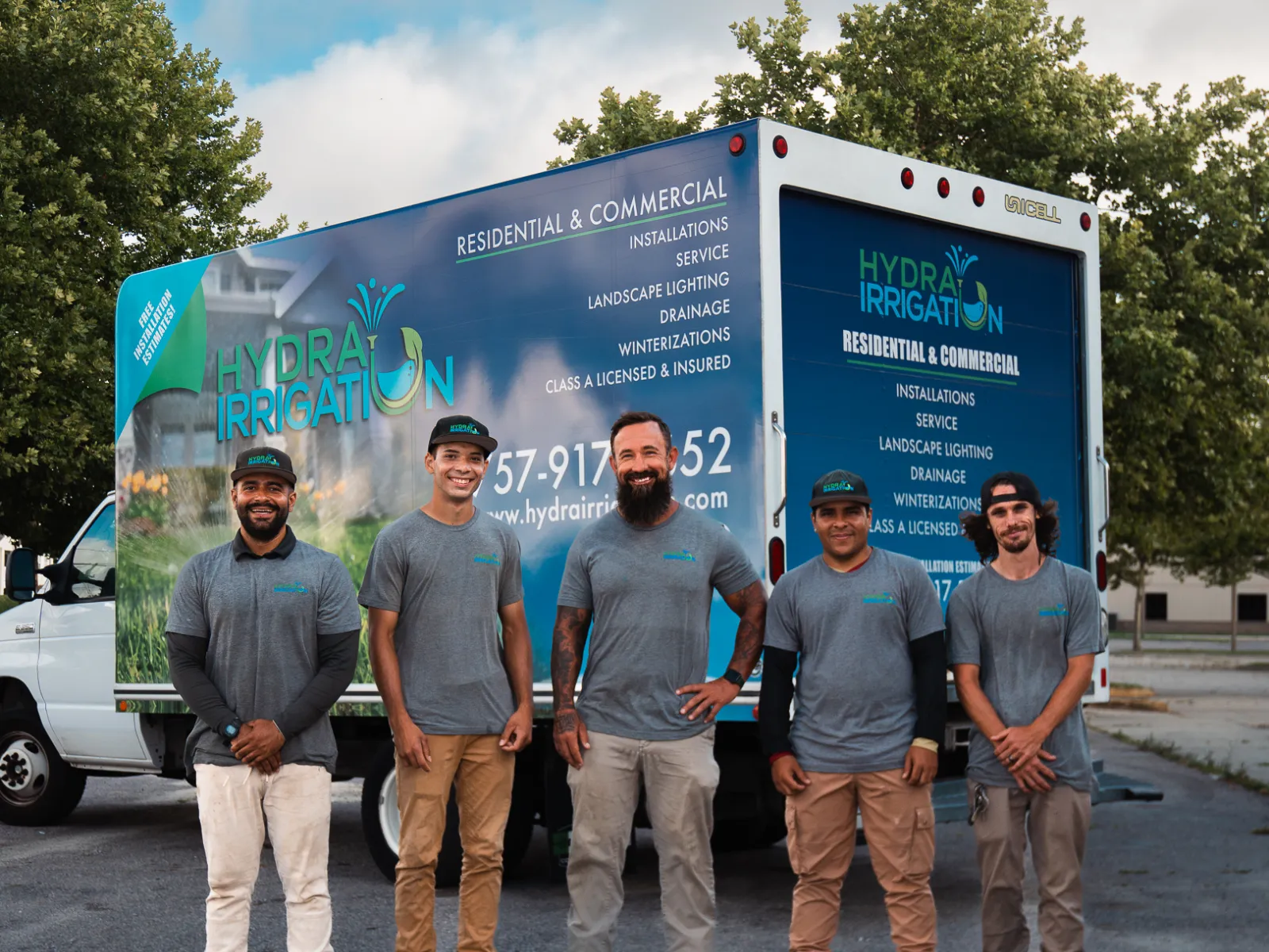 Five men in grey shirts and work pants stand in front of a Hydra Irrigation service truck with trees in background.