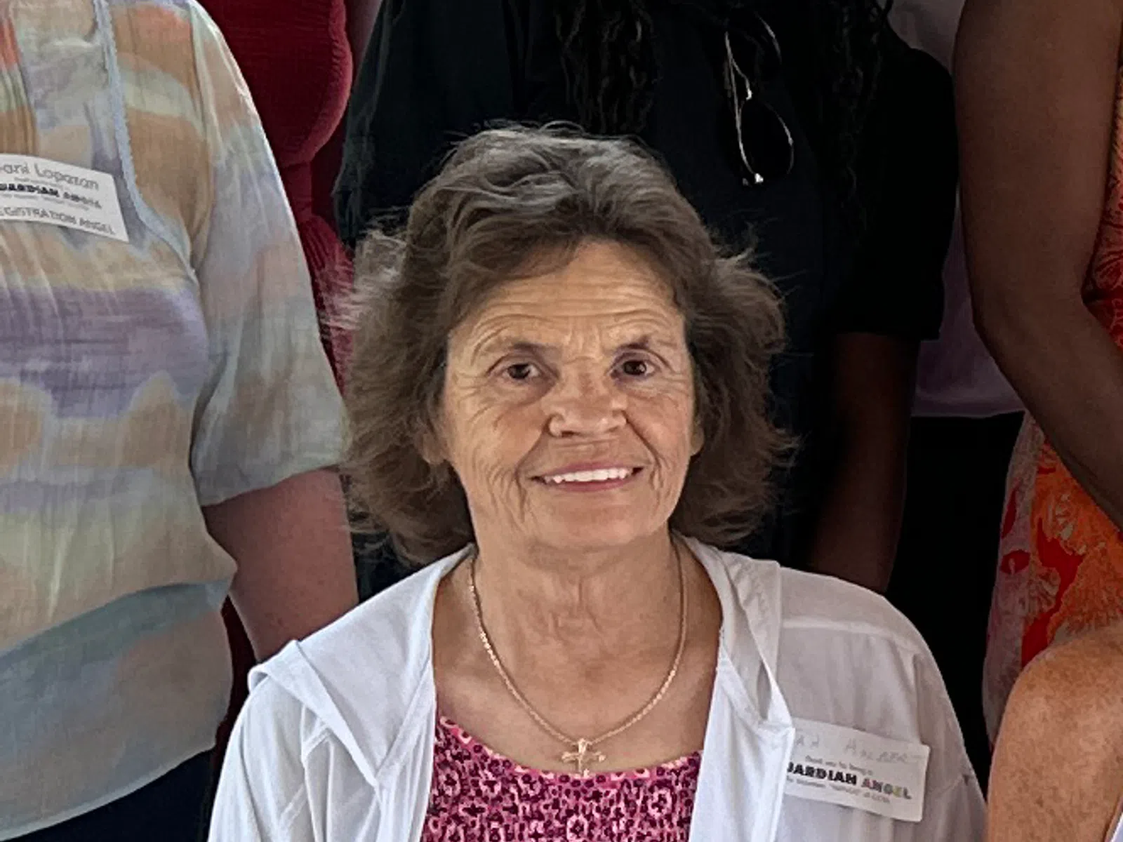 Elderly woman smiling, wearing a pink patterned dress and white jacket, surrounded by other people indoors.