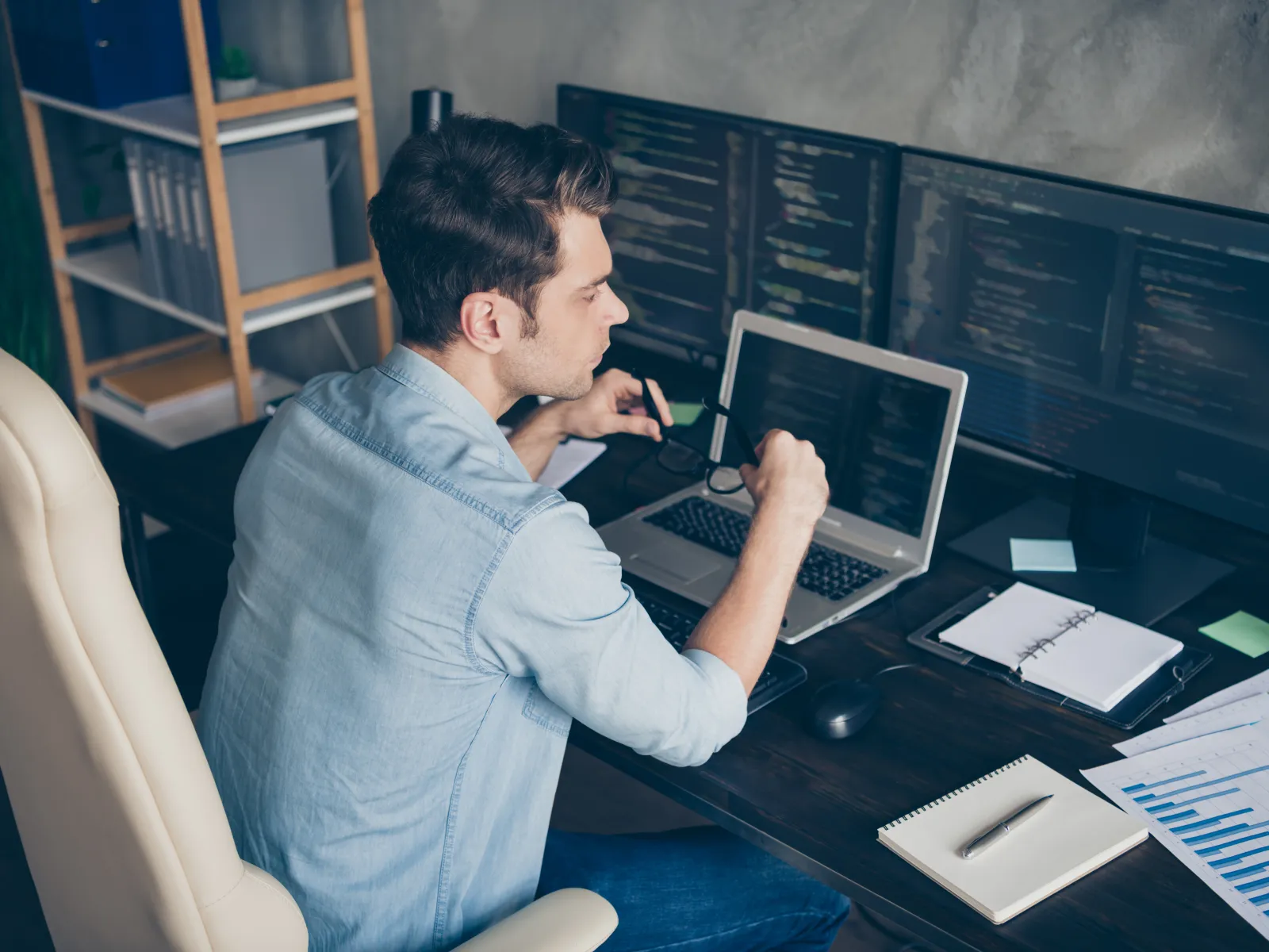 Young man analyzing code on laptop and desktop monitors while working at a home office desk.