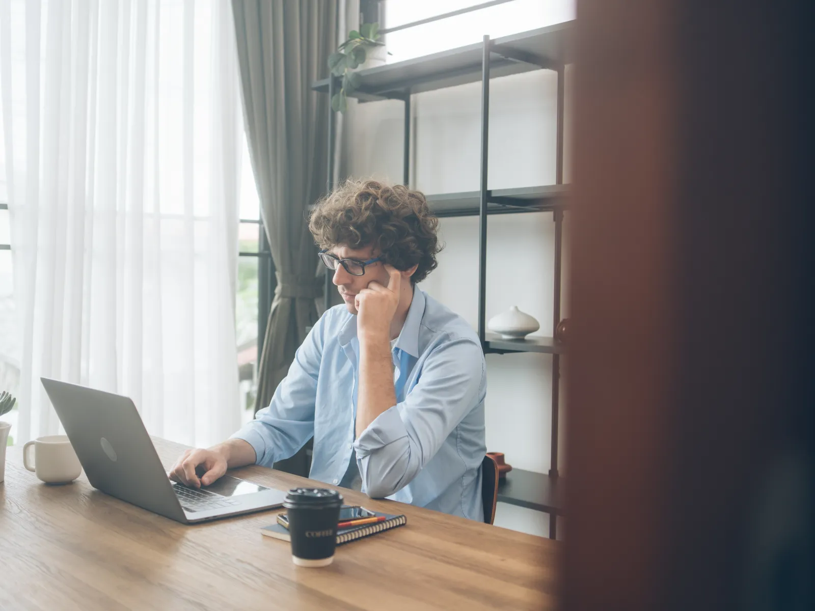 Man with glasses working on laptop at wooden desk with coffee cup and notebook in bright modern room