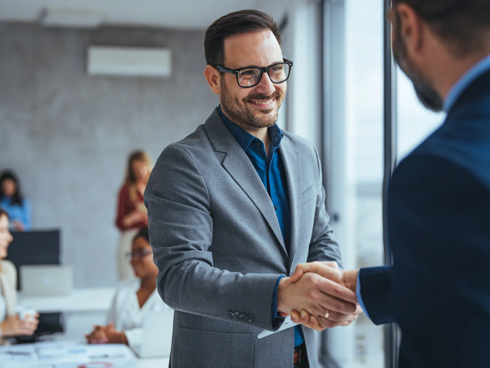 Two businessmen shaking hands in a modern office with colleagues working in the background