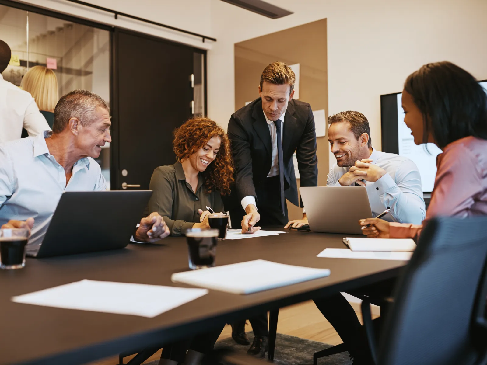 Group of professionals collaborating in a modern meeting room around a table with laptops and drinks.