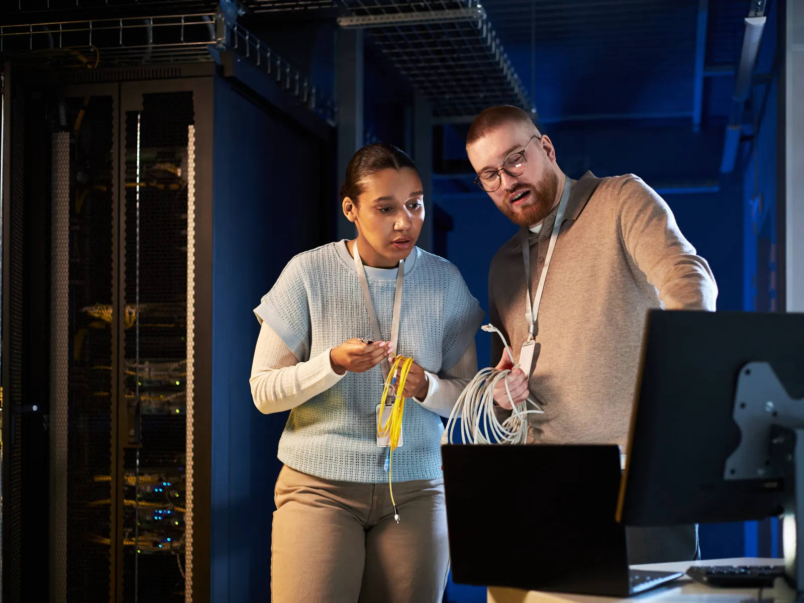 Two IT technicians inspecting network cables and working on a laptop in a server room with racks.