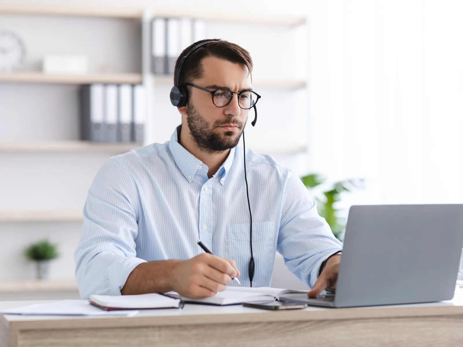 Focused man wearing headset working on laptop and taking notes in a bright office environment.