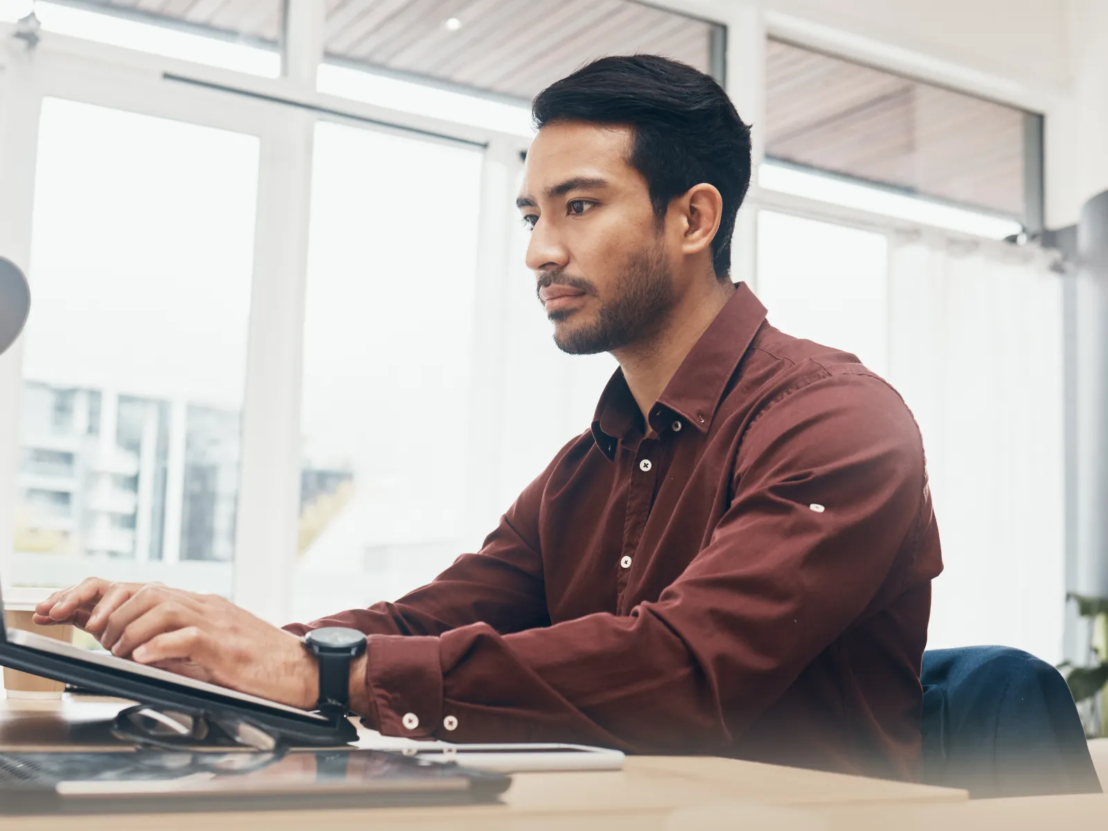 Young man focused on his laptop working at a bright office desk with plants and stationery nearby.