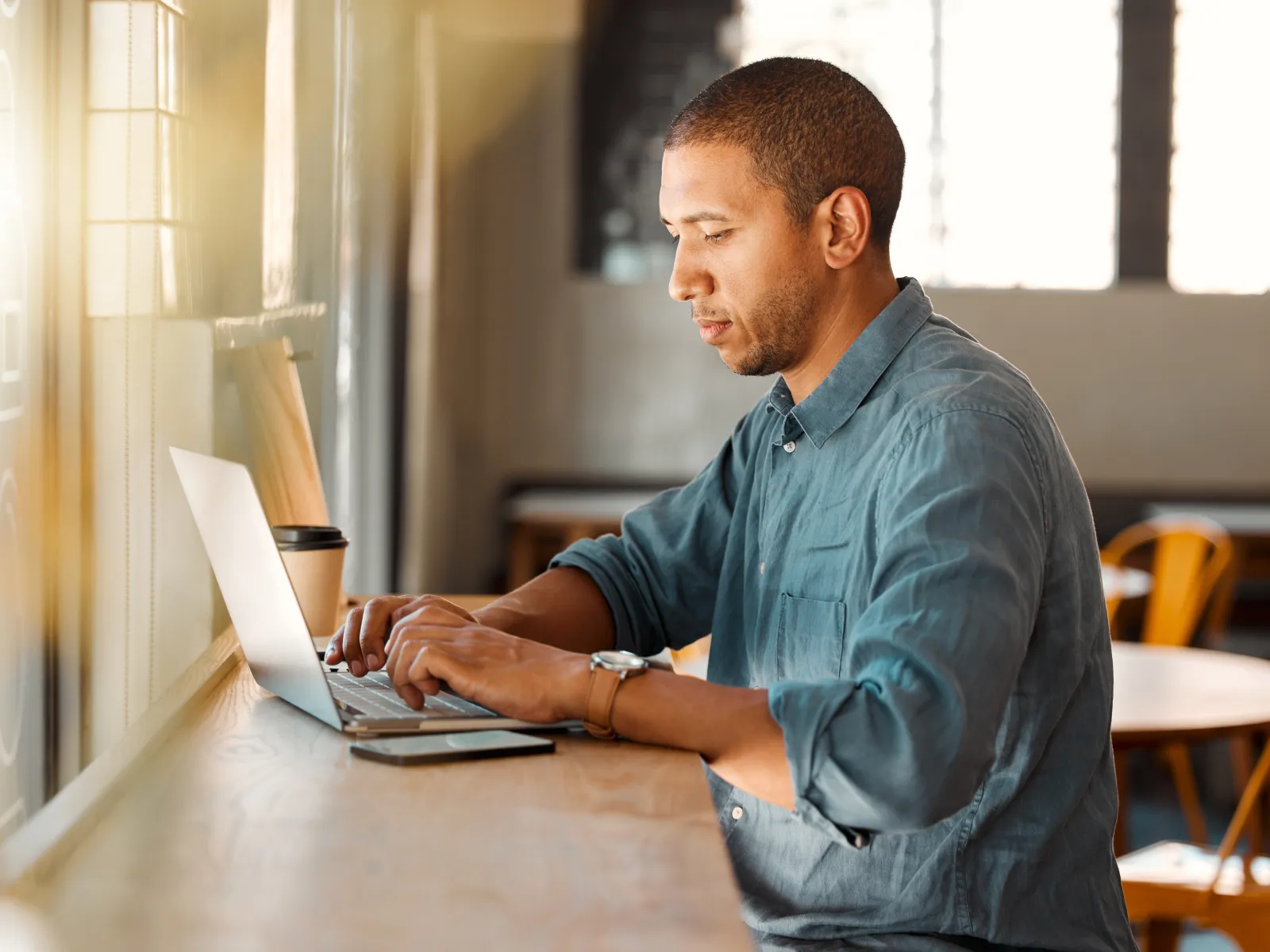 Man working on laptop at a wooden desk next to a window with a coffee cup nearby in a sunlit cafe.