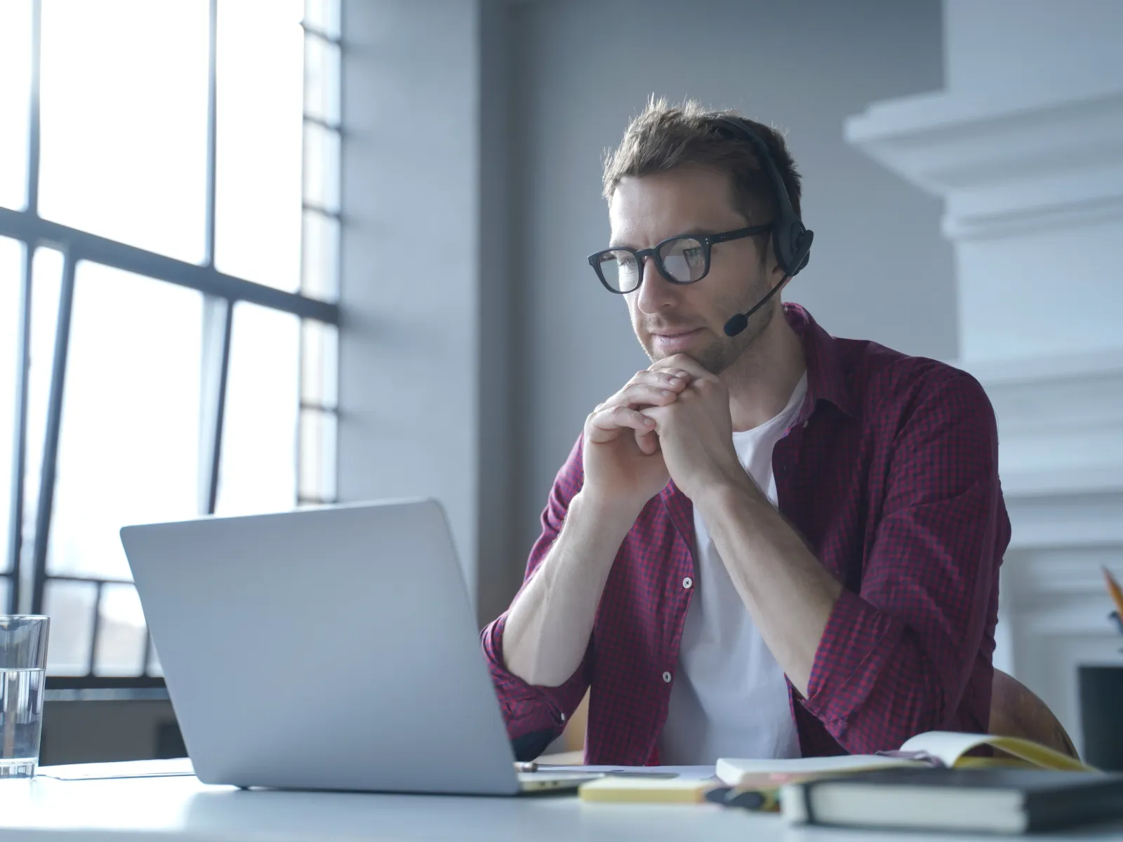 Man wearing glasses and headset working on a laptop in a bright modern office with books and a glass of water.