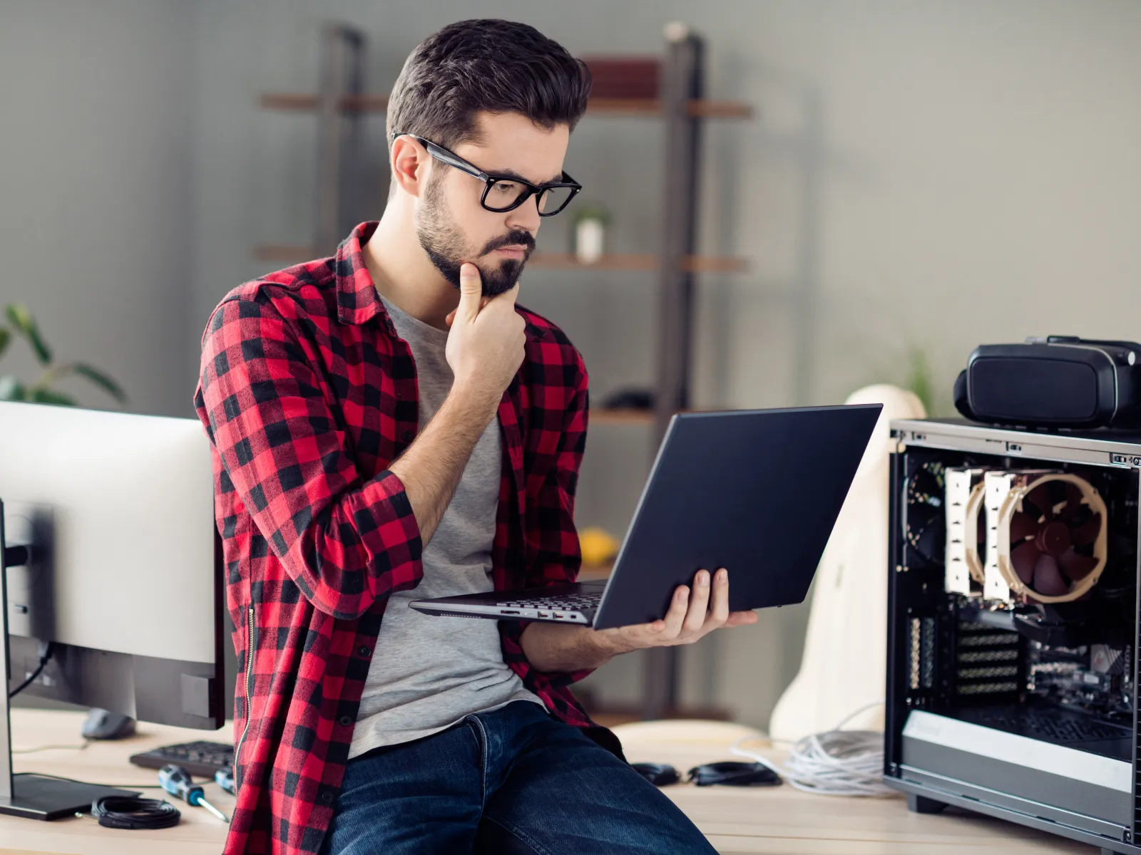 Man in glasses and red plaid shirt working on laptop next to an open desktop computer in a modern office.