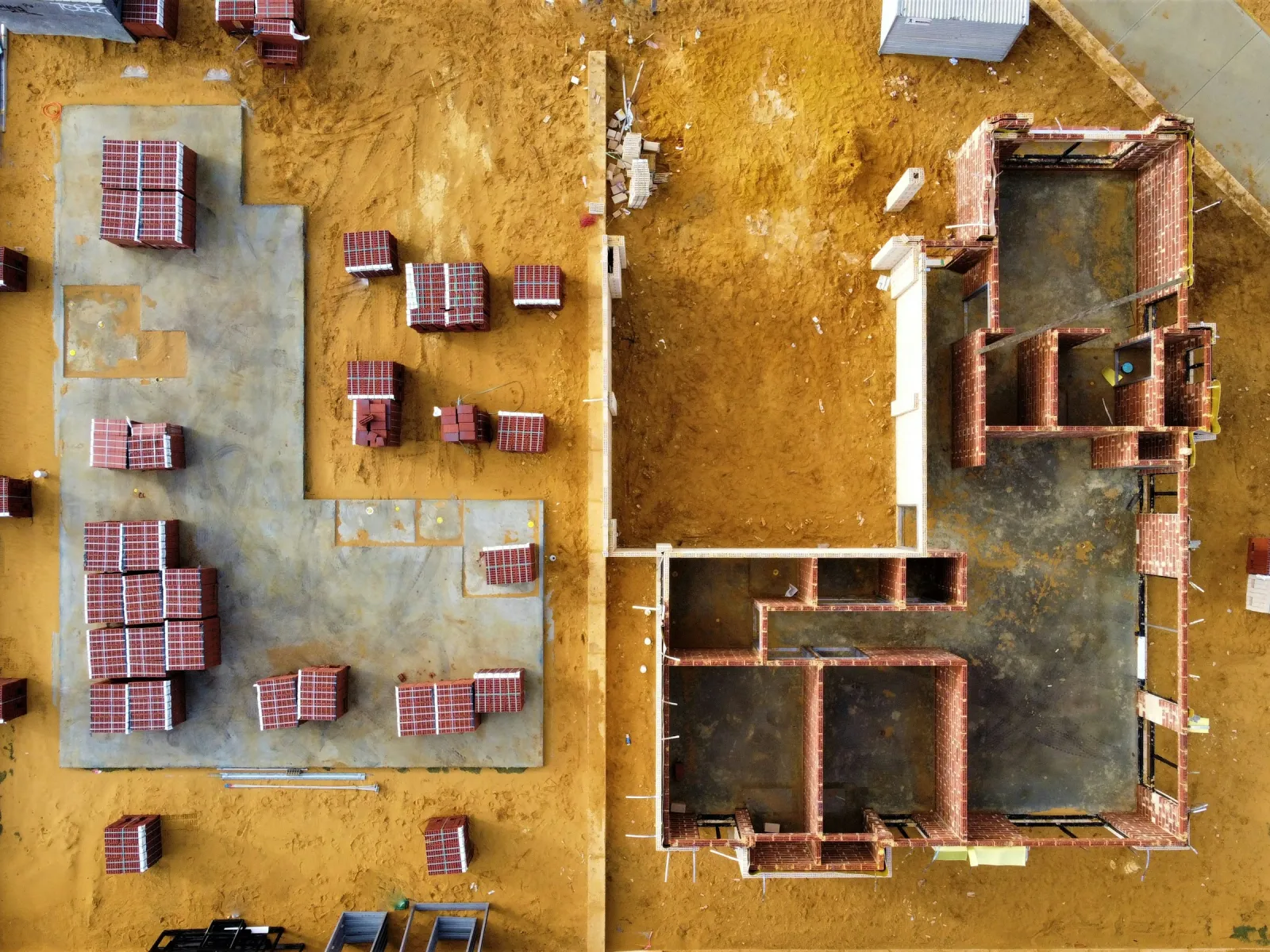 Aerial view of a construction site showing house foundations and stacks of bricks on sandy ground.