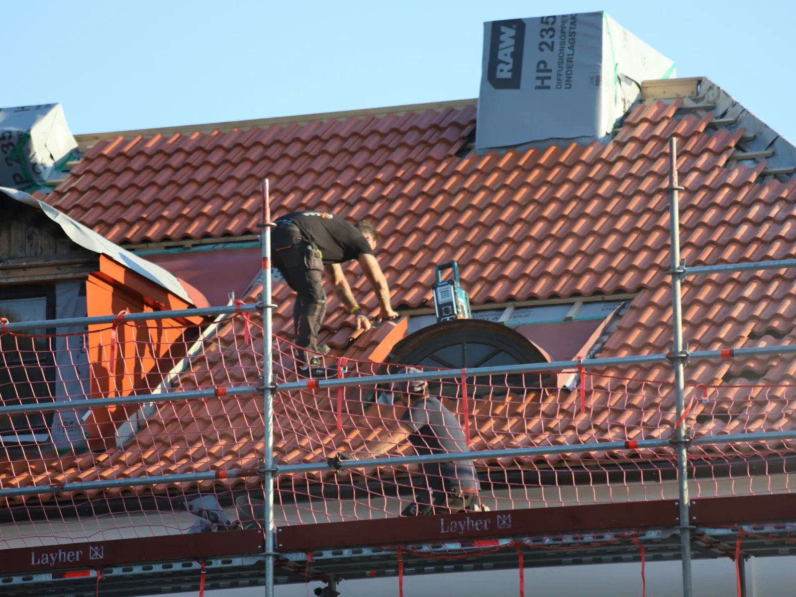 Two workers installing red clay roof tiles on a house under blue sky with safety scaffolding in place.