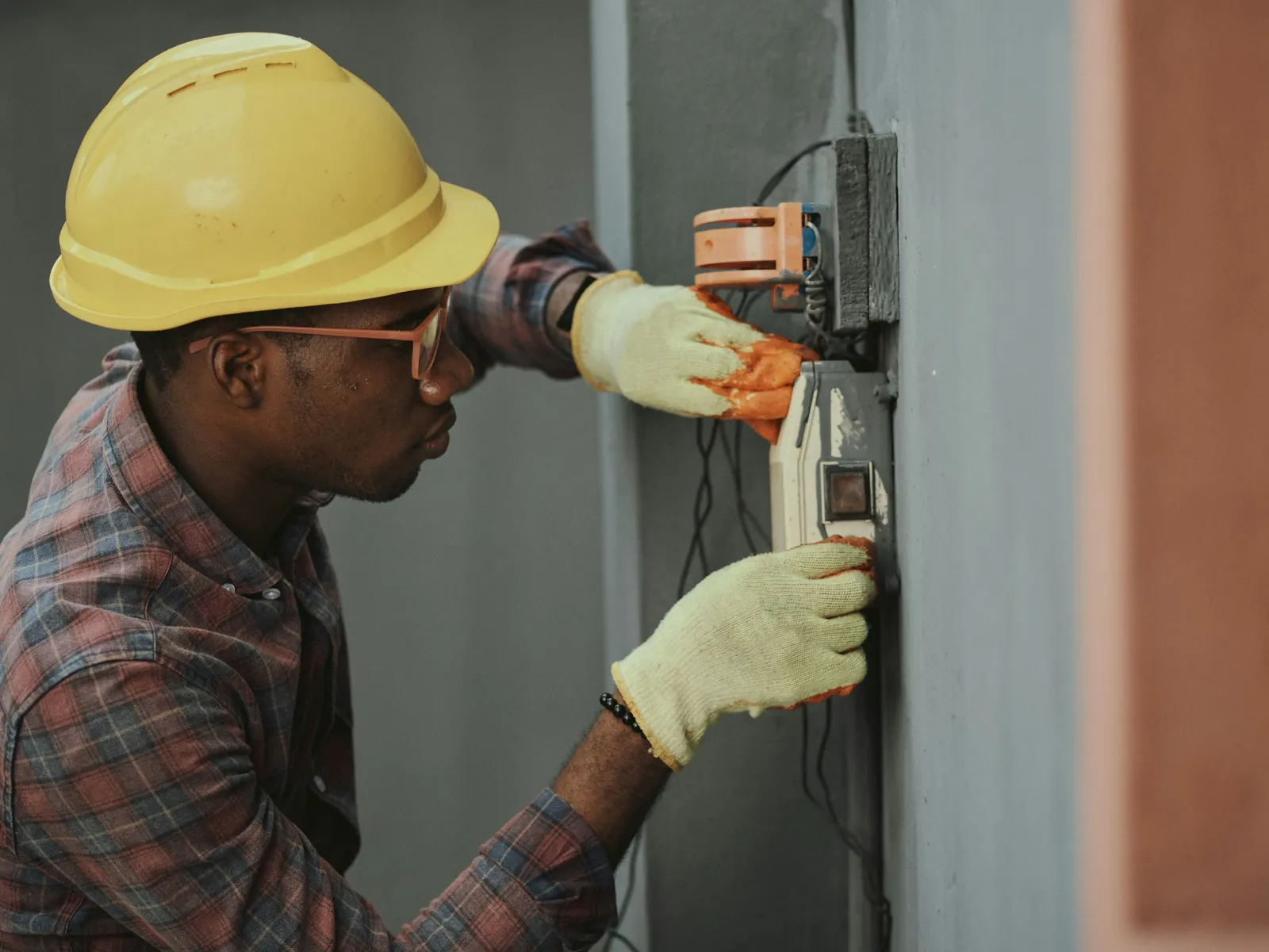 Electrician wearing a yellow helmet and gloves working on an electrical meter mounted on a wall indoors.